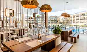 Ravintola, jossa puiset pöydät ja vaaleat tuolit, koristeltu kasveilla ja lampuilla, näkymä uima-altaalle ja hotellirakennuksiin. (Tekoälyn luoma teksti)