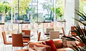 Valoisa ja moderni istuma-alue, jossa on pöytiä ja tuoleja, sekä suuret ikkunat, jotka tarjoavat näkymän ulkona olevaan vehreyteen. (Tekoälyn luoma teksti)