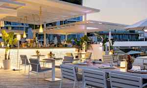 Ulkoterassi, jossa on valkoisia kalusteita ja vihreitä kasveja, lähellä lasi-ikkunoilla varustettua rakennusta. (Tekoälyn luoma teksti)