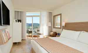 Hotellihuone, jossa on suuri sänky, oleskelualue ja uloskäynti parvekkeelle, josta on näkymä puihin ja maisemaan. (Tekoälyn luoma teksti)