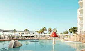 Kuva uima-altaasta, jossa on kotilon muotoinen liukumäki ja sienimäinen vesiputous. Taustalla näkyy aurinkotuoleja ja varjoja. (Tekoälyn luoma teksti)