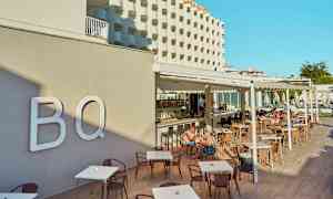 Hotellin julkisivu ja ulkoterassi, jolla on pöytiä ja tuoleja, ihmisiä syömässä ja istumassa. (Tekoälyn luoma teksti)