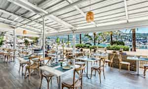 Ulkoterassi meren rannalla, jossa on pöytiä ja tuoleja sekä näkymä satamassa oleviin veneisiin. (Tekoälyn luoma teksti)