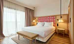 Hotellihuone, jossa on parivuode, vaaleat verhot ja penkki sängyn edessä. (Tekoälyn luoma teksti)