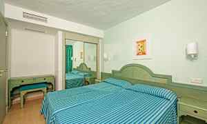 Hotellihuone, jossa on kaksi erillistä vuodetta, sinivihreäraitaista vuodevaatetta, kirjoituspöytä jakkaroineen ja peilikaappi. (Tekoälyn luoma teksti)