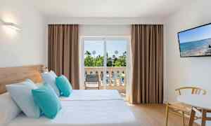 Hotellihuone, jossa on kaksi yksittäisiä vuodetta vaaleansinisillä ja turkooseilla tyynyillä, parveke näkymällä puutarhaan sekä seinällä oleva TV. (Tekoälyn luoma teksti)