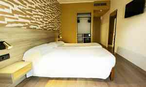 Hotellihuone, jossa kaksi yhden hengen vuodetta, seinään kiinnitetty televisio ja vaatekaappi. (Tekoälyn luoma teksti)