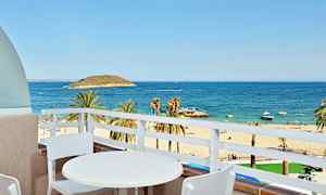 Näköala parvekkeelta, jossa on valkoinen pöytä ja kaksi tuolia, hiekkarannalle, palmuihin ja siniselle merelle. (Tekoälyn luoma teksti)