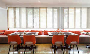 Huone, jossa on pöytiä ja tuoleja, koristeltu oransseilla tyynyillä ja aurinkosuojaisilla ikkunoilla. (Tekoälyn luoma teksti)