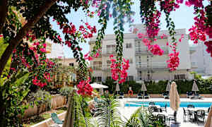 Rakennus, jossa on useita parvekkeita, riippuvien vaaleanpunaisten kukkien takana, uima-allas ja aurinkotuoleja. (Tekoälyn luoma teksti)