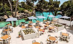 Terassi, jossa on pöytiä, tuoleja ja aurinkovarjoja lähellä turkoosia vesirantaa ja vehreyttä. (Tekoälyn luoma teksti)