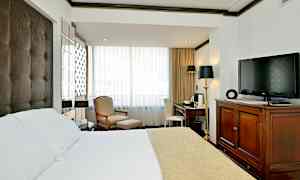 Hotellihuone, jossa on sänky valkoisella ja beigenvärisellä päiväpeitteellä, nojatuoli ja jalkatuki ikkunan vieressä, kirjoituspöytä tuoleineen sekä televisio puukomuutin päällä. (Tekoälyn luoma teksti)