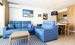 Olohuone, jossa sinisiä sohvia, sohvapöytä, seinälle ripustettuja tauluja ja keittiö, jossa puisia yksityiskohtia taustalla. (Tekoälyn luoma teksti)