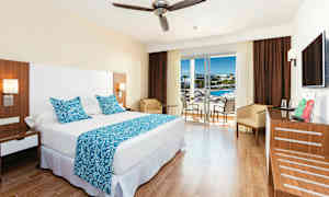 Hotellihuone, jossa suuri sänky, siniset koristeelliset tyynyt ja peite sekä pöytä tuoleineen lasioven vieressä, joka johtaa parvekkeelle. (Tekoälyn luoma teksti)