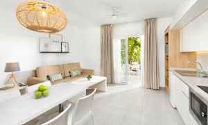 Vaalea ja modernisti sisustettu huone, jossa beige sohva, valkoinen ruokapöytä ja keittiö sekä avonaiset ovet kalustetulle parvekkeelle. (Tekoälyn luoma teksti)