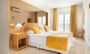 Hotellihuone, jossa on parivuode, valkoiset lakanat ja keltaiset tyynyt ja päiväpeite. Huoneessa on vaaleat puuhuonekalut ja ikkunat verhoineen. (Tekoälyn luoma teksti)