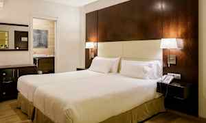 Hotellihuone, jossa on suuri sänky, kaksi yöpöytälamppua, yöpöytä ja puhelin. Taustalla näkyy kylpyhuoneen oviaukko. (Tekoälyn luoma teksti)