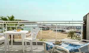 Näkymä parvekkeelta, jolla on valkoiset ulkokalusteet, näkymänä ranta, pienvenesatama ja meri taustalla. (Tekoälyn luoma teksti)