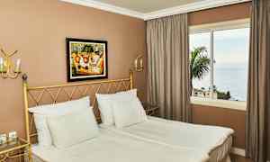 Kuva hotellihuoneesta, jossa on kaksi sänkyä, taulu seinällä ja ikkunasta merinäkymä. (Tekoälyn luoma teksti)