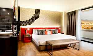 Hotellihuone, jossa on suuri sänky, moderni sisustus ja istuinpenkki sängyn edessä. Ikkunasta näkyy maisema vuorille ja rakennuksille. (Tekoälyn luoma teksti)