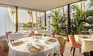 Ravintola, jossa on katettuja pöytiä, suuret ikkunat ja näkymä palmuihin. (Tekoälyn luoma teksti)