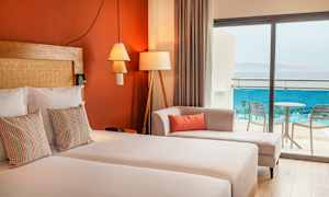 Hotellihuone, jossa on parivuode, koristeellisia tyynyjä, sohva ja lasiovet, jotka johtavat parvekkeelle merinäkymällä. (Tekoälyn luoma teksti)