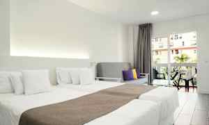 Hotellihuone, jossa on kaksi erillistä sänkyä valkoisin vuodevaattein, harmaa sohva värillisillä tyynyillä ja parveke näkymällä. (Tekoälyn luoma teksti)