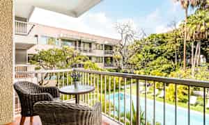 Näkymä parvekkeelta, jolla on kaksi rottinkituolia pienen pöydän ääressä, uima-allas ja viheralueita sekä asuinrakennus taustalla. (Tekoälyn luoma teksti)