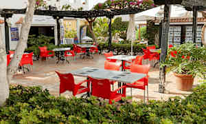 Ulkoruokailutila, jossa on punaisia tuoleja ja pöytiä, ympäröitynä kasvillisuudella. (Tekoälyn luoma teksti)