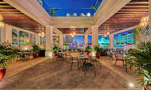 Ulkotarjoilualue, jossa pöytiä ja tuoleja katon alla, ympäröity palmuilla ja valaistuksella. (Tekoälyn luoma teksti)