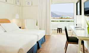 Valoisa ja moderni hotellihuone, jossa on kaksi erillistä vuodetta, valkoinen nojatuoli ja työpöytä lampulla. Suuri ikkuna johtaa parvekkeelle, jolta on näkymä vuorille ja vehreyteen. (Tekoälyn luoma teksti)