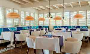 Tyylikäs ravintola, jossa on valkoiset pöytäliinat, siniset sisustuselementit ja modernit kattovalaisimet. (Tekoälyn luoma teksti)