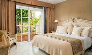 Hotellihuone, jossa on suuri sänky, beiget vuodevaatteet, valkoiset tyynyt ja lasiovi, joka avautuu terassille, jolla on vihreää kasvillisuutta. (Tekoälyn luoma teksti)