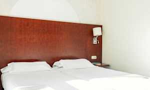 Kaksi erillissänkyä, joissa valkoiset vuodevaatteet ja tumma puinen sängynpääty. Seinälamppu on kiinnitetty sivulle. (Tekoälyn luoma teksti)