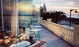 Pariskunta syö illallista lasiseinän lähellä, näkymä terassille ja merelle. (Tekoälyn luoma teksti)