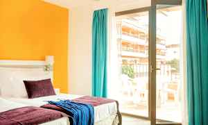 Hotellihuone, jossa on sänky, värikkäitä tyynyjä ja tekstiilejä sekä lasiovi, joka johtaa parvekkeelle. (Tekoälyn luoma teksti)