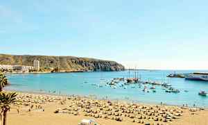 Ranta aurinkotuoleilla ja varjoilla suojaisan merenlahden vieressä, taustalla näkyy veneitä ja rakennuksia. (Tekoälyn luoma teksti)