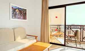 Näkymä hotellihuoneesta, jossa on beigesoja, puupöytä ja parveke, jolta näkyy ranta. (Tekoälyn luoma teksti)