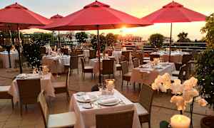 Ulkoterassi, jossa punaisia aurinkovarjoja, katettuja pöytiä ja auringonlasku taustalla. (Tekoälyn luoma teksti)