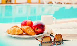 Lautanen, jossa on croissanteja ja omenoita, sijaitsee pöydällä kirjan ja aurinkolasien vieressä. Taustalla näkyy uima-allas. (Tekoälyn luoma teksti)