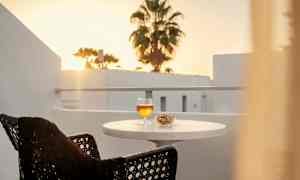 Parveke, jossa pöydällä on juomalasi ja kulho pähkinöitä. Taustalla näkyy auringonlasku ja palmu. (Tekoälyn luoma teksti)