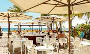 Ulkoilmaravintola, jossa valkoiset pöydät ja tuolit varjojen alla, näkymä merelle. (Tekoälyn luoma teksti)