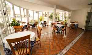 Ruokailutila, jossa on useita pöytiä ja tuoleja, ympäröitynä ikkunoilla, joiden näkymä on puutarhaan. (Tekoälyn luoma teksti)