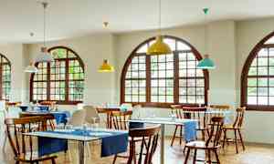 Ravintola, jossa puupöydät ja tuolit, siniset pöytäliinat ja kaarevakehyksiset ikkunat. (Tekoälyn luoma teksti)