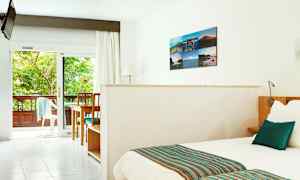 Valoisa hotellihuone, jossa sänky, pöytä ja tuolit sekä näkymä vehreälle parvekkeelle. (Tekoälyn luoma teksti)