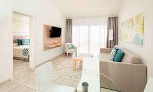 Kuvassa on valoisa ja moderni olohuone, jossa on sohva, televisio, ruokapöytä sekä tuoleja. Ovi johtaa makuuhuoneeseen. (Tekoälyn luoma teksti)