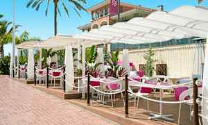 Ulkoravintola, jossa on valkoiset varjot ja pöydillä vaaleanpunaiset liinat. Palmuja ja rakennus taustalla. (Tekoälyn luoma teksti)