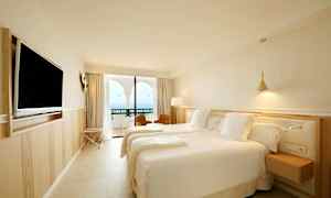 Hotellihuone, jossa on kaksi sänkyä, seinälle kiinnitetty televisio ja parveke merinäköalalla. (Tekoälyn luoma teksti)