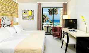 Hotellihuone, jossa on sänky, tuoli, kirjoituspöytä ja televisio. Lasiseinän takana on parveke merinäköalalla. (Tekoälyn luoma teksti)