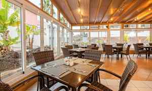 Ruokasali, jossa rottinkituoleja ja puupöytiä, ympäröity suurilla lasi-ikkunoilla, joista näkymä puutarhaan. (Tekoälyn luoma teksti)